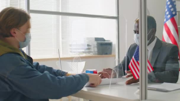 ガラスシールドの後ろに机に座って保護面マスクのアフリカ系アメリカ人領事館やコロナウイルスパンデミック中の米国大使館で働くビザ申請者 — ストック動画