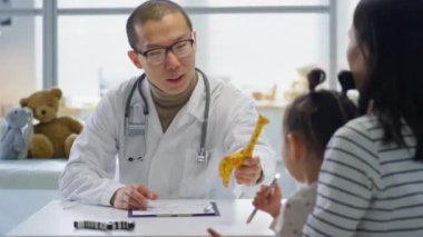Beyaz önlüklü ve gözlüklü Asyalı bir çocuk doktoru küçük şirin bir kıza zürafa oyuncağı veriyor ve klinikte danışmanlık yaparken annesiyle konuşuyor.