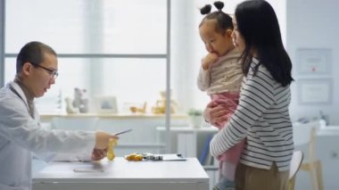 Asyalı anne küçük kızıyla klinikte yürüyor, erkek çocuk doktoruna tıbbi geçmişi veriyor ve onunla konuşuyor.