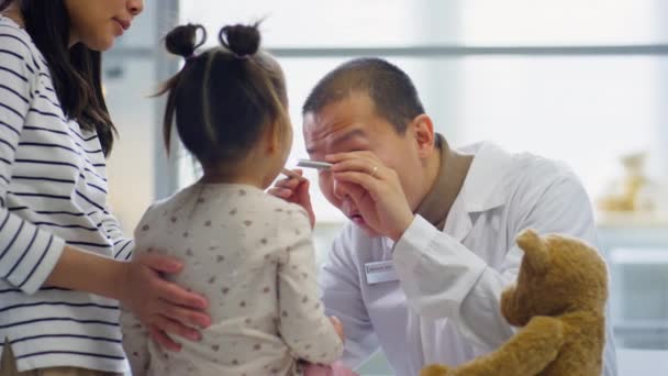 Azjatycki Pediatra Badający Gardło Dziewczynki Latarką Podczas Gdy Matka Bawi — Wideo stockowe