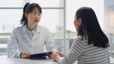 Beyaz önlüklü Asyalı kadın doktor tıbbi geçmişi tutuyor ve klinikte çalışırken kadınlara danışmanlık yapıyor.