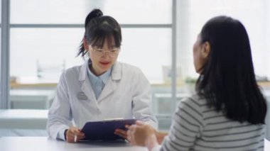 Beyaz önlüklü ve gözlüklü Asyalı kadın doktor klinikte çalışırken masada oturuyor ve kadınlara tıbbi danışmanlık yapıyor.