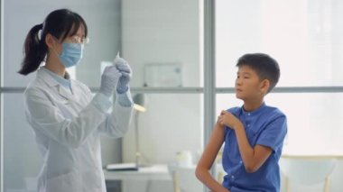 Koruyucu maskeli ve steril eldivenli kadın doktor klinikteki Asyalı ergen çocuğa anti-covid aşı enjekte ediyor.