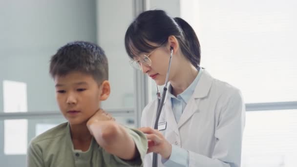 아시아 의사가 아이를 청진기로 진료소에서 검진을 엄지손가락을 그에게 — 비디오