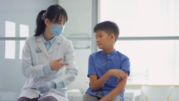 클리닉에 아시아인 소년에게 접종을 준비하는 방법을 설명하는 마스크를 여의사 — 비디오