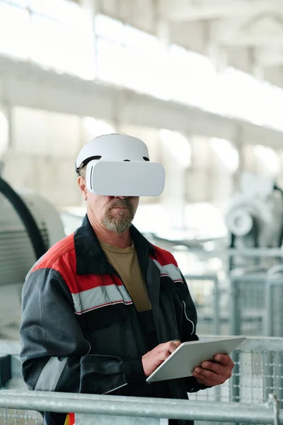 Vertical image of mature engineer using digital tablet and wearing VR glasses to watch project online while working in factory