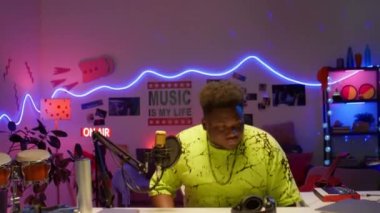 Afrikalı Amerikalı müzik yapımcısı renkli neon ışıklarla ses stüdyosunda yürüyor, masada oturuyor, dizüstünü açıyor, kulaklıkları takıyor ve işe hazırlanırken mikrofonu ayarlıyor.