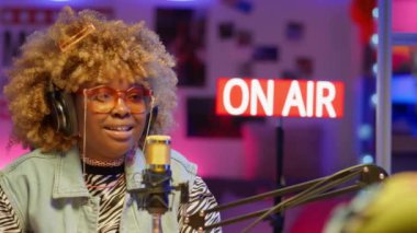 Moda güneş gözlüklü Afrikalı Amerikalı bir kadının, stüdyoda, neon ışıklı bir konukla podcast yaparken mikrofonla konuşurken çekilen omuz üstünden.