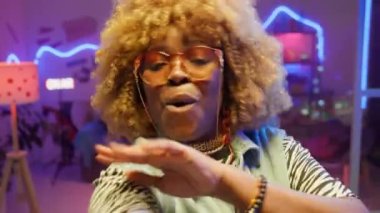 Moda kıyafetli ve güneş gözlüklü mutlu Afro-Amerikan kadın POV 'u. Sosyal medya için müzik videosu çekerken kameraya bakıyor ve neon ışıkla dans ediyor.