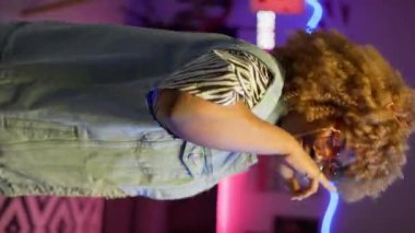 Renkli neon ışıklarıyla aydınlatılmış ve stüdyo arkadaşlarıyla eğlenen, dans eden, gözlüklü Afrikalı genç bir kadının dikey çekimi.