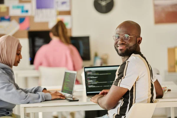 非裔美国人程序员一边和计算机坐在工作场所 一边和同事一起工作 一边对着相机微笑 — 图库照片