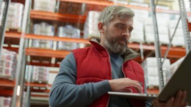 Orta yaşlı müdürün iş günü depoda durup dizüstü bilgisayarda yazı yazışının düşük açılı görüntüsü.