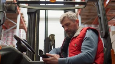 Depoda çalışırken akıllı telefon kullanan gri saçlı ve sakallı orta yaşlı bir forklift operatörü.
