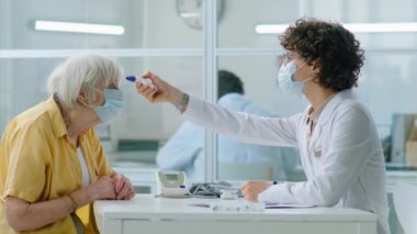 Korunaklı yüz maskesi takan kadın doktor klinikte koronavirüs kontrolü sırasında ateşini ölçerken kızılötesi termometresi olan yaşlı bir kadının alnını tarıyor.