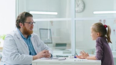 Erkek çocuk doktoru ve küçük kız klinikte masada oturuyor ve tıbbi danışmanlık sırasında konuşuyorlar.