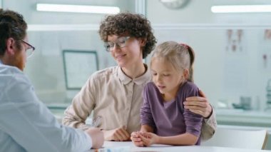 Anne, küçük neşeli kızını kucaklıyor ve muayenehanede erkek çocuk doktoruyla konuşuyor.