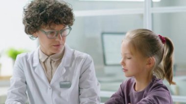 Beyaz önlüklü ve gözlüklü kadın çocuk doktoru klinikte çalışırken küçük güzel bir kızla konuşuyor.