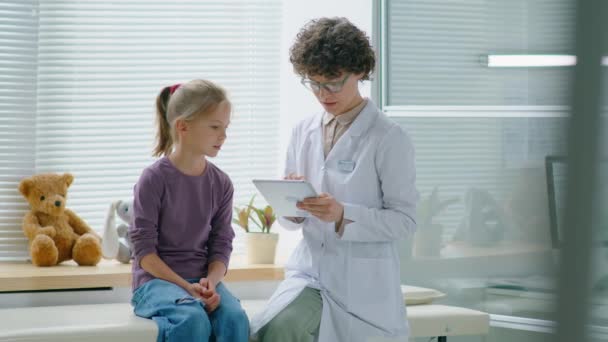 診療所で診察中に医務室で一緒に座っている間 女性医師はデジタルタブレットを使用し 女の子と話をします — ストック動画