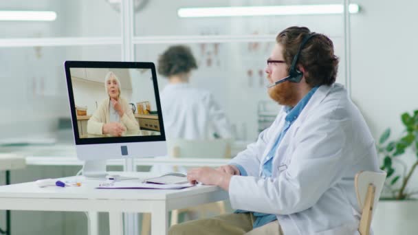 医務室からのオンライン健康相談を与えながら コンピュータ上のオンラインビデオ通話を介して高齢女性と話してヘッドセットの男性医師 — ストック動画