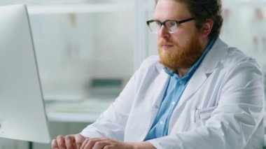 Tıp bürosunda masa başında çalışırken, sakallı, kablosuz bilgisayar klavyesine yazı yazan genç bir doktor.