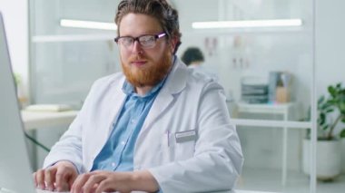 Gözlüklü ve beyaz ceketli genç sakallı doktorun portresi klinikteki masada otururken kameraya gülümseyerek poz veriyor.