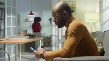 Kadın mutfakta bir şeyler yaparken Afro-Amerikan bir adamın evdeki kanepeye oturup akıllı telefondan mesaj yazışının yan görüntüsü.