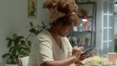 Genç Afrikalı Amerikalı bir kadının evde yemek masasında otururken akıllı telefonuyla yazışının arkası.
