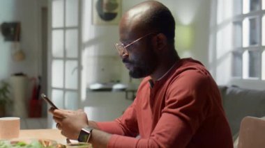 Afrika kökenli Amerikalı bir adamın cep telefonuyla mesajlaşırken evdeki yemek masasında otururken ki görüntüsü.