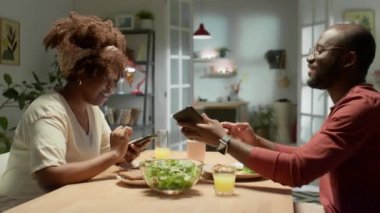 Genç Afro-Amerikan çiftin akıllı telefonlarda internette gezinirken ve sosyal medya paylaşımlarını tartışırken evde yemek yerken.