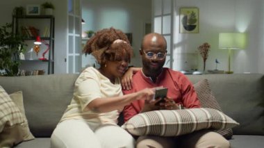 Afro-Amerikalı çiftin gülümseyip, akıllı telefondaki sosyal medya paylaşımlarını tartışırken akşamları evdeki kanepede dinlenirken görüntüsünü yakınlaştırın.