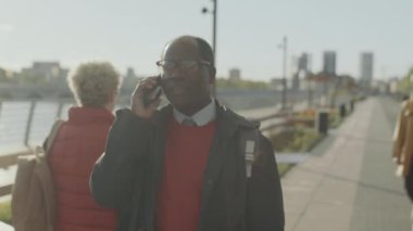 Sırt çantalı Afrikalı Amerikalı işadamının belden yukarı vuruşu cep telefonuyla konuşurken kenar mahallelerde yürürken.