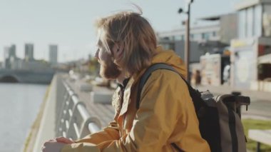 Sırt çantalı genç sakallı adam nehir manzarasını seyrederken şehir setinde duruyor ve korkuluklara yaslanıyor.