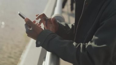 Nehir kenarındaki korkuluklara yaslanan ve akıllı telefondan mesaj yazan Afro-Amerikan adamı yakın çekim.