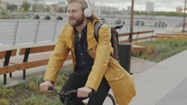 Kablosuz kulaklıklı genç beyaz adam, şehirdeki nehir kenarında bisiklet sürüyor ve gülümsüyor.