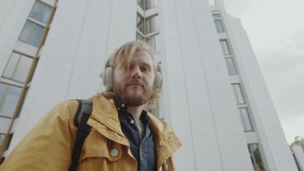 髪の長い若い髭の男の低角度の肖像画は ワイヤレスヘッドフォンで音楽を聞いて 街の路上でカメラのポーズ — ストック動画