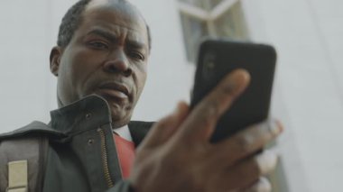 Orta yaşlı Afro-Amerikan iş adamının şehir caddesinde dikilip akıllı telefon kullanmasının düşük açılı görüntüsü.
