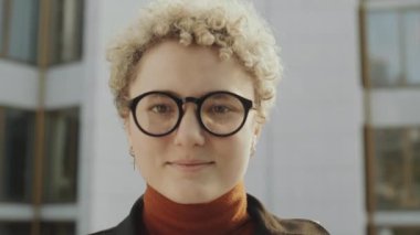 Sarışın, kıvırcık saçlı, gözlüklü bir kızın portresi şehir sokaklarında durup kameraya gülümseyerek poz veriyor.