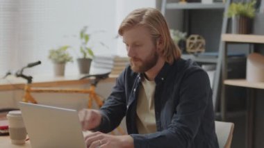 Uzun saçlı genç sakallı iş adamı dizüstü bilgisayarla çalışıyor ve ofiste çalışırken kameraya poz veriyor.