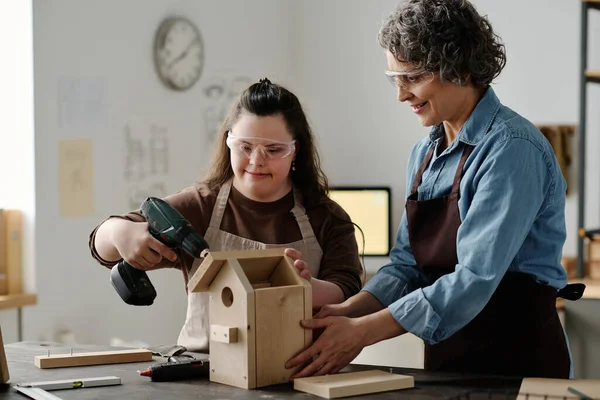 Girl with down syndrome using drill to make birdhouse with woman helping her with it in workshop