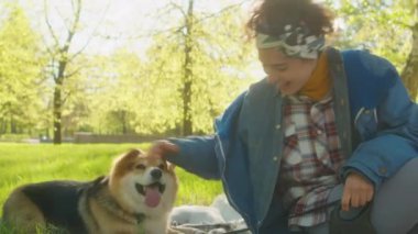 Yeşil çimlerde battaniye üzerinde oturan ve parkta gününü geçirirken oynak Corgi köpeğini okşayan bir kadın.