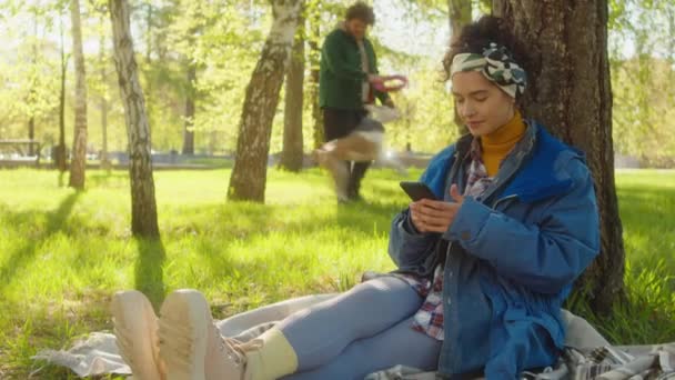 年轻女子坐在公园里一棵树旁的草地上 用智能手机和爱抚可爱的科奇犬四处走动 — 图库视频影像