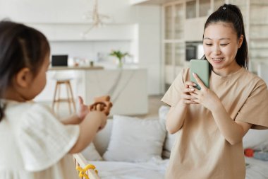 Mutlu Asyalı anne, onlar odada oynarken çocuğunun akıllı telefonuyla fotoğrafını çekiyor.