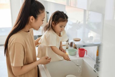 Genç anne kızına mutfakta bulaşık yıkamayı öğretiyor.