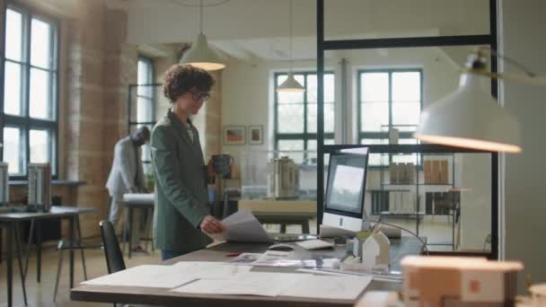 短い巻き毛を持つ女性建築家は オフィスの職場に歩いて 机に座って ドキュメントをチェックし マグカップからコーヒーを飲む — ストック動画