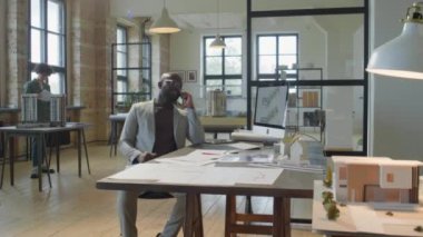 Afrika kökenli Amerikalı işadamının ofis masasında otururken görüntüsünü yakınlaştırın ve iş günü mimarlık ofisinde cep telefonuyla konuşun.