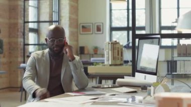 Cep telefonuyla konuşan ve açık uzay ofisindeki masada çalışırken not alan Afrikalı Amerikalı erkek mimar.