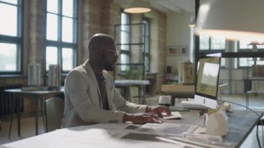 Bilgisayarda çalışan neşeli Afro-Amerikan mimarın portresi ve sonra ofiste gülümseyen bir şekilde kameraya poz vermek.