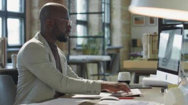 Ofiste çalışırken bilgisayarda yazan ve kağıtları kontrol eden Afrikalı Amerikalı mimar.