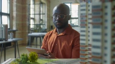 Afrika kökenli Amerikalı erkek mimarın dijital tablet kullanırken ve emlak projesinde çalışırken konut binalarının modelini incelerken.