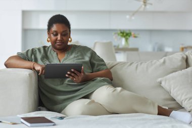 Afro-Amerikalı kadın oturma odasında kanepede oturuyor ve dijital tablet üzerinde çalışıyor.